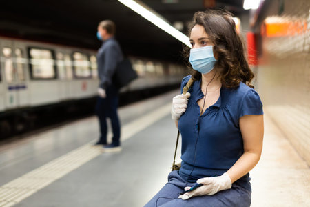 Young woman in mask and protective gloves waiting for subway train at underground station, enjoying favorite music on earphones. Concept of prevention and social distancing in coronavirus pandemicの写真素材