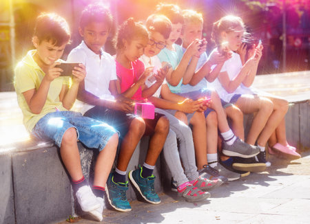 Group of smiling children sitting at urban street with mobile devices