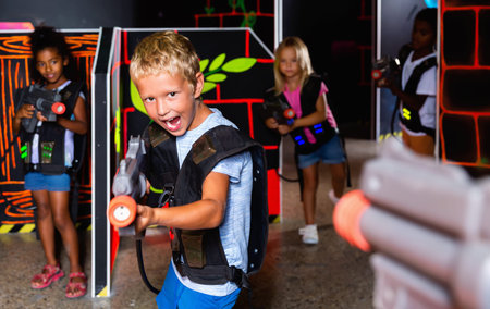 Excited little cheerful positive boy aiming laser gun at other players during lasertag game in dark roomの写真素材