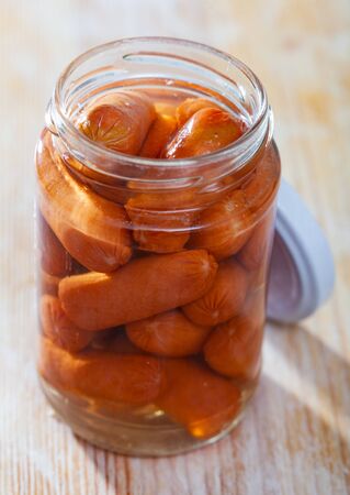 Open jar with appetizing preserved mini sausages on wooden tableの写真素材