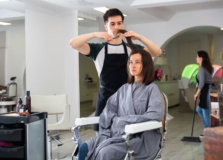 Positive male hairdresser making hair styling for young woman, working day in beauty salonの写真素材