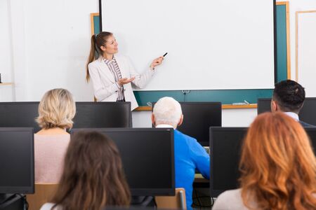 Friendly young female teacher lecturing attentive adult students of different ages in computer roomの写真素材