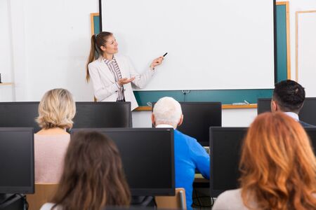 Friendly young female teacher lecturing attentive adult students of different ages in computer room