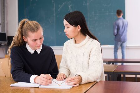 Intelligent girl student helping her Chinese girl schoolmate prepare for exam in classroomの写真素材