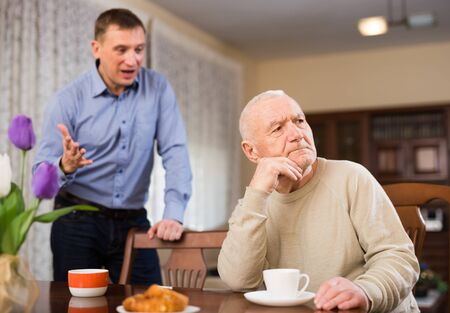 Offended elderly man sitting at table with disgruntled adult son standing behind and reprimanding himの写真素材
