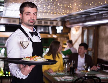 Man waiter is holding tray with wine and salad for clients in restaurant.の写真素材
