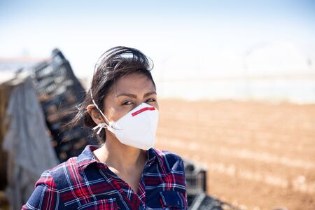 Portrait of adult Hispanic female farmer in medical face mask standing on farm field on sunny spring day. Concept of dust protection or coronavirus infection prevention during workの写真素材