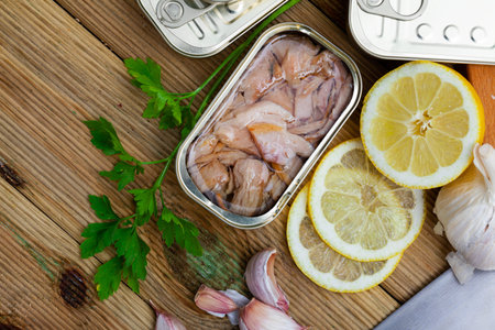 Appetizing smoked tuna belly slices in oil in open tin can on wooden table with greens and lemon....