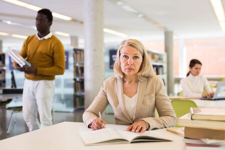 Elderly woman makes notes in notebook and read book in public libraryの写真素材