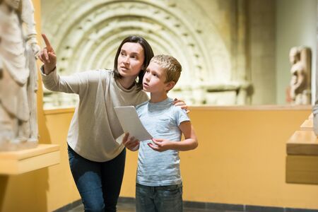 Young woman with son observing with interest sculptures exhibition in art museum, pointing to something interestingの写真素材