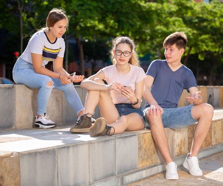 Teenagers having friendly discussion and using cellphones during gathering outdoors on summer dayの写真素材