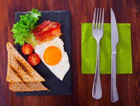 Picture of fried eggs with   bacon, toasted bread, tomatoes and lettuce at plate on tableの写真素材