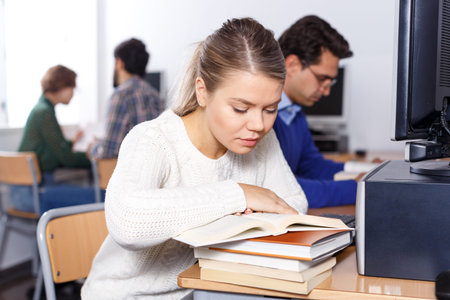 Attractive girl preparing for exam in college library, reading book and using pc for searching...