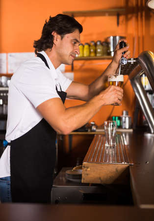 Smiling man barman pouring golden beer to glass in cafeの写真素材