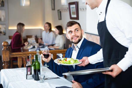 Waiter serving delicious salads to handsome young man at restaurantの写真素材