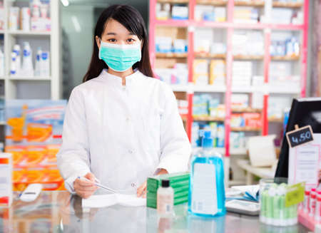 Chinese woman pharmacist in protective facial mask keeps track of drugs in interior of pharmacyの写真素材