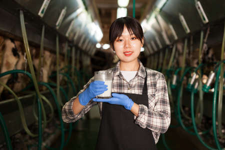 Milkmaid in apron standing with glass of fresh milk at farm in cowshedの写真素材