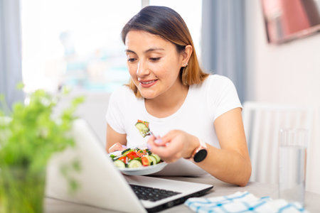 Cheerful female who is working with laptop at the table at the home and eating saladの写真素材