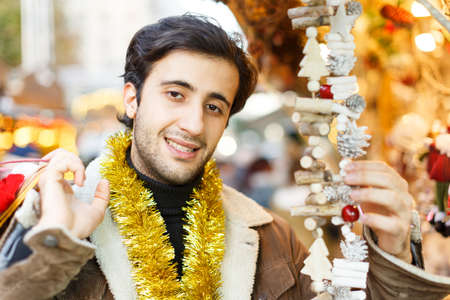 Happy young man with garland looking Christmas toys at fair outdoorの写真素材