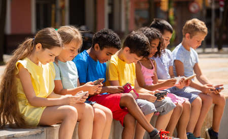 Positive children with mobile devices sitting at urban streetの写真素材