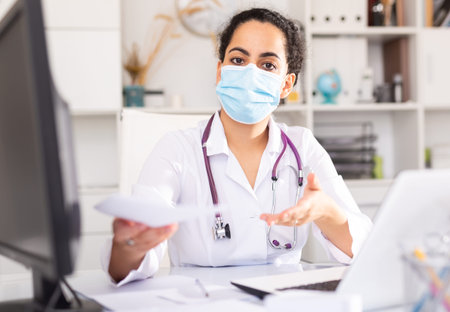 Female therapist in protective mask working at table with laptop in office