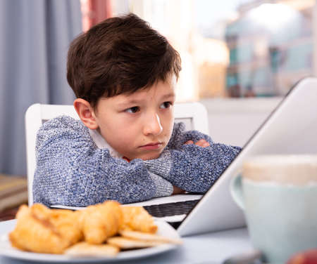 Sad boy with laptop having breakfast in the kitchenの写真素材
