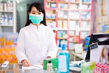 Chinese woman pharmacist in protective facial mask keeps track of drugs in interior of pharmacyの写真素材