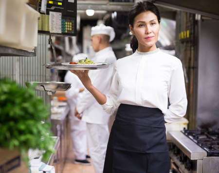 portrait of happy female waitress standing in white kitchen in cafeの写真素材