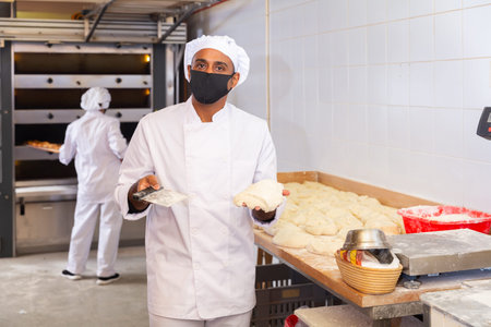 Bakery worker in protective mask cuts raw dough with knife to prepare for bakingの写真素材