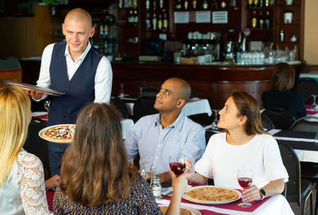 Man waiter is brings pizza to friends who have dinner and drink wineの写真素材
