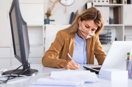 Business woman in protective medical mask is working with project behind laptopの写真素材