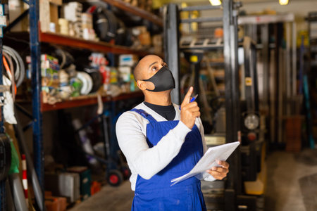 Warehouse worker in protective mask making notes during inventory of building materials