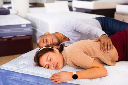 Young latin american couple sleeping on display sample bed with orthopedic mattress in furniture shopの写真素材
