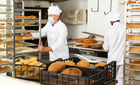 Man in protective mask transporting cart with bread in bakery