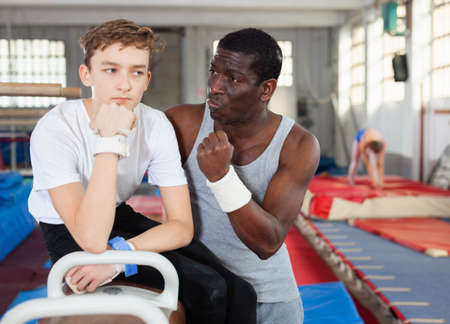 Male coach talking with upset teenage boy on gymnastic equipment at acrobatic centerの写真素材