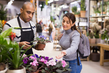 Shop assistant helping woman to choose geranium in flower shop