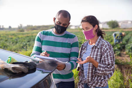 Hispanic farmer in medical mask discussing papers with female workmate near carの写真素材