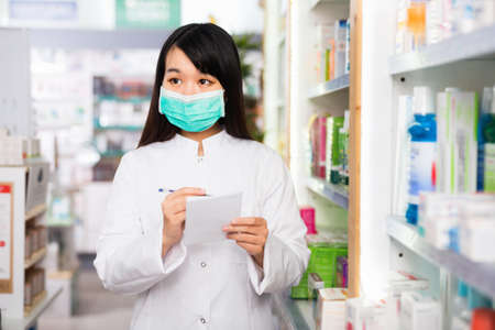 Chinese woman pharmacist in protective facial mask keeps track of drugs in interior of pharmacyの写真素材