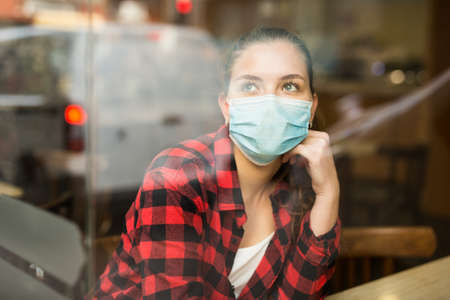 Portrait of a thoughtful girl in a protective mask, sitting at a table in a cafeの写真素材