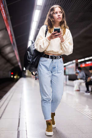 Thoughtful girl, walking along the platform of a subway station, holds a mobile phoneの写真素材