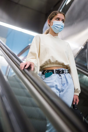 Focused girl in a protective mask in the subway goes down the escalator