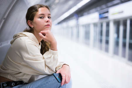 Portrait of a thoughtful girl sitting on a bench at a metro station waiting for a trainの写真素材