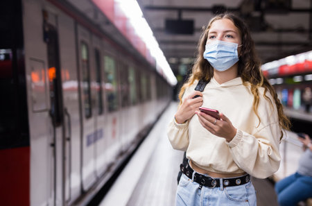 Young woman in mask using mobile phone while waiting metro