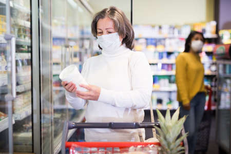 Mature woman in protective mask choosing food products on shelves in shopの写真素材