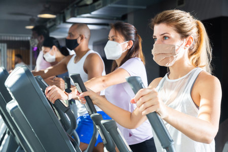 Slim athletic people in protective masks running on treadmill in fitness club