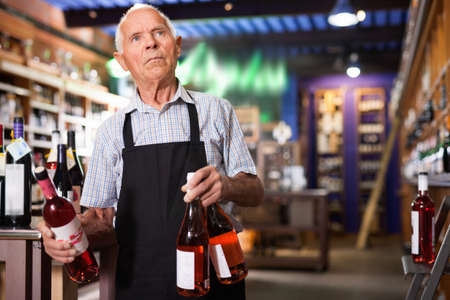 Confident elderly salesman of wine house arranging wine bottles on shelves rackの写真素材