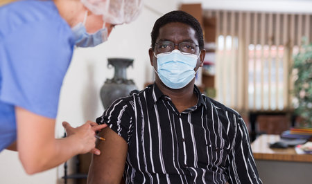 Male nurse injecting vaccine in shoulder of male patient during covid vaccination in office