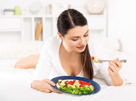 Pretty woman in white dress eating vegetable salad in bedの写真素材