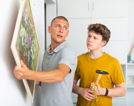 Father shows his son how to hang painting on the wall of house
