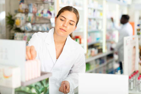 Portrait of a young female pharmacist laying out goods on the shelves of a rackの写真素材